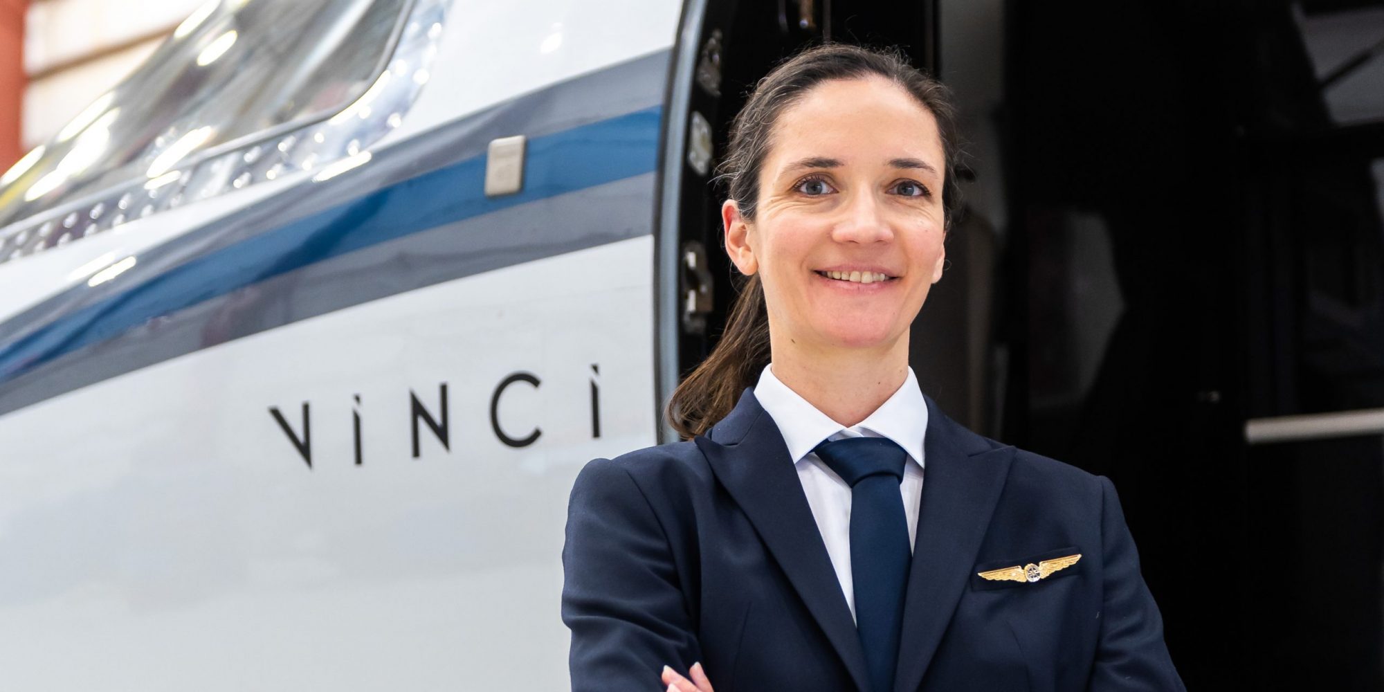 Une pilote souriante se tient devant un avion, portant une tenue professionnelle avec une cravate et une épingle de pilote. Le nom "Vinci" est visible sur l'appareil.