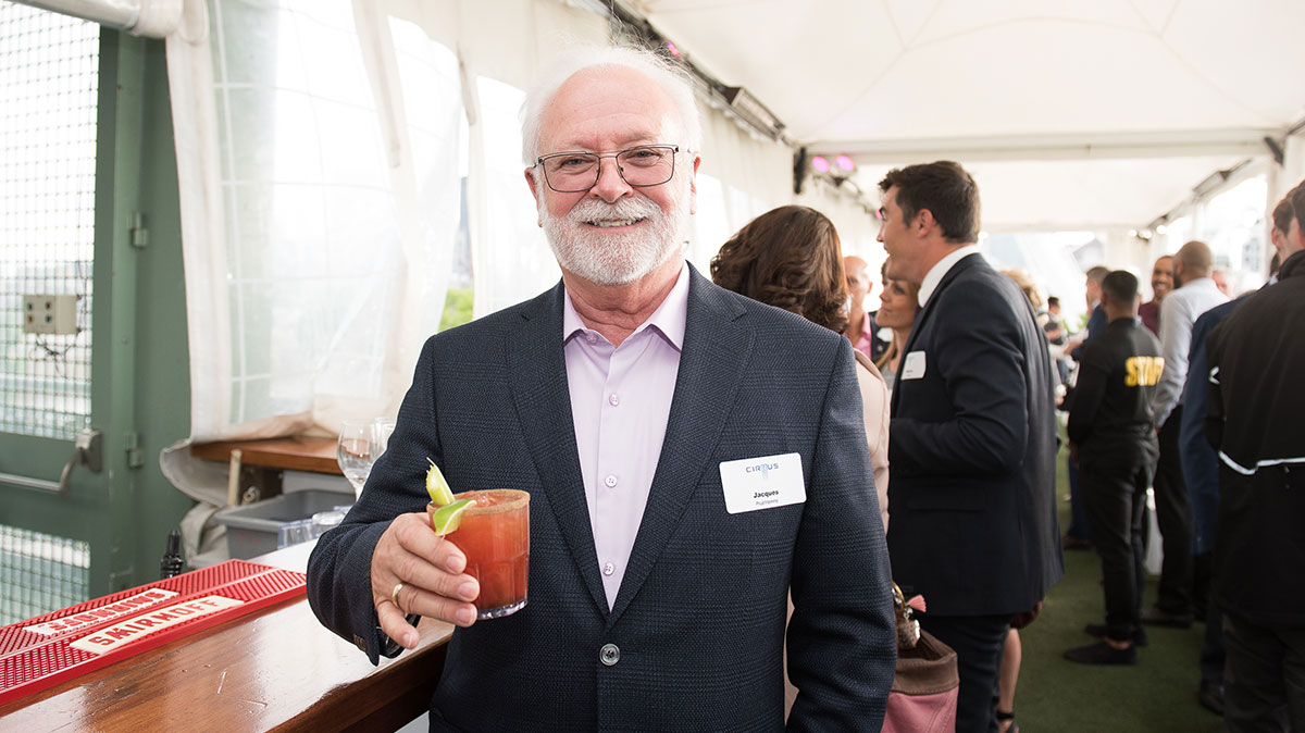Un homme souriant tient un cocktail dans un événement en plein air, entouré de personnes en conversation. Une ambiance conviviale règne.