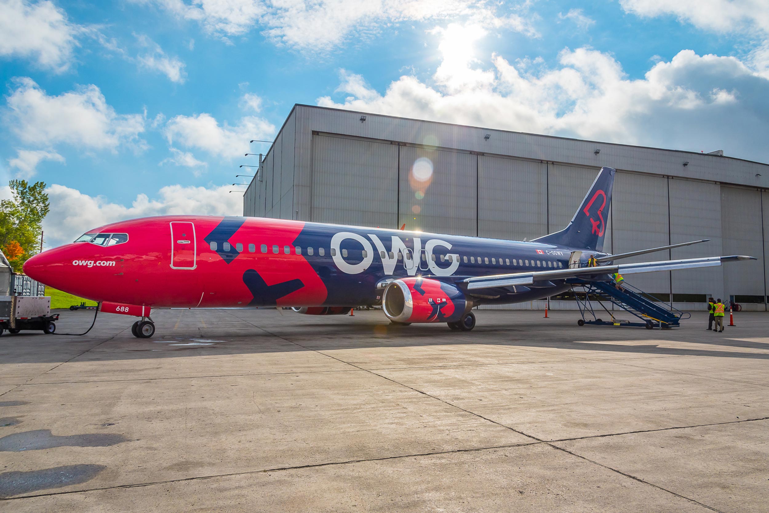 A brightly colored OWG airplane parked outside a hangar, with a boarding staircase and ground crew nearby under a partly cloudy sky.