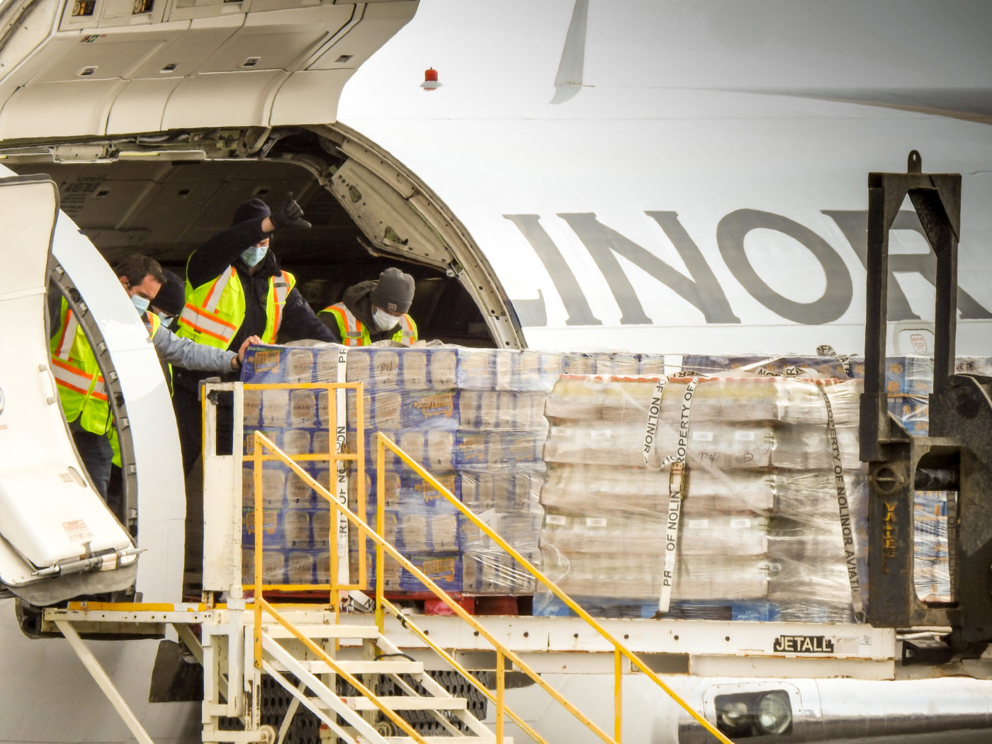 Des travailleurs en gilets de sécurité déchargent des palettes de marchandises d'un avion cargo, portant des masques.