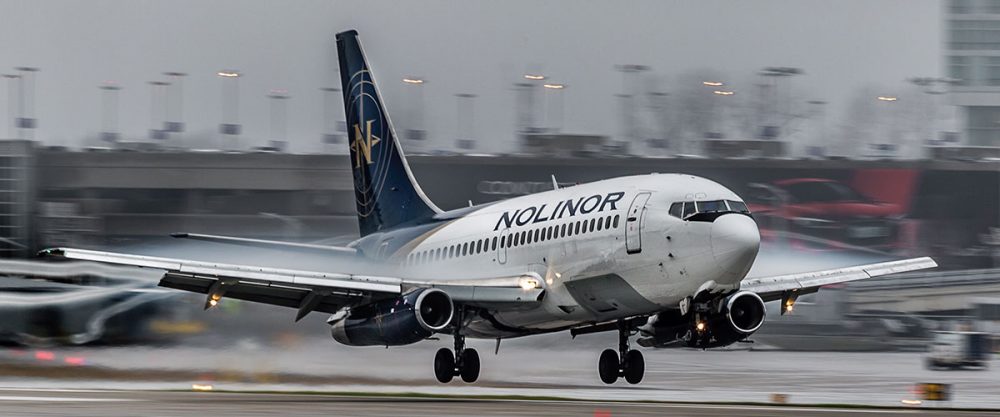Un avion de la compagnie Nolinor atterrit sur une piste mouillée, avec des lumières d'aéroport en arrière-plan. Le ciel est nuageux.