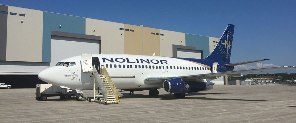 A Nolinor Aviation aircraft parked at a hangar, with stairs leading up to the entrance, showcasing its blue and white livery.