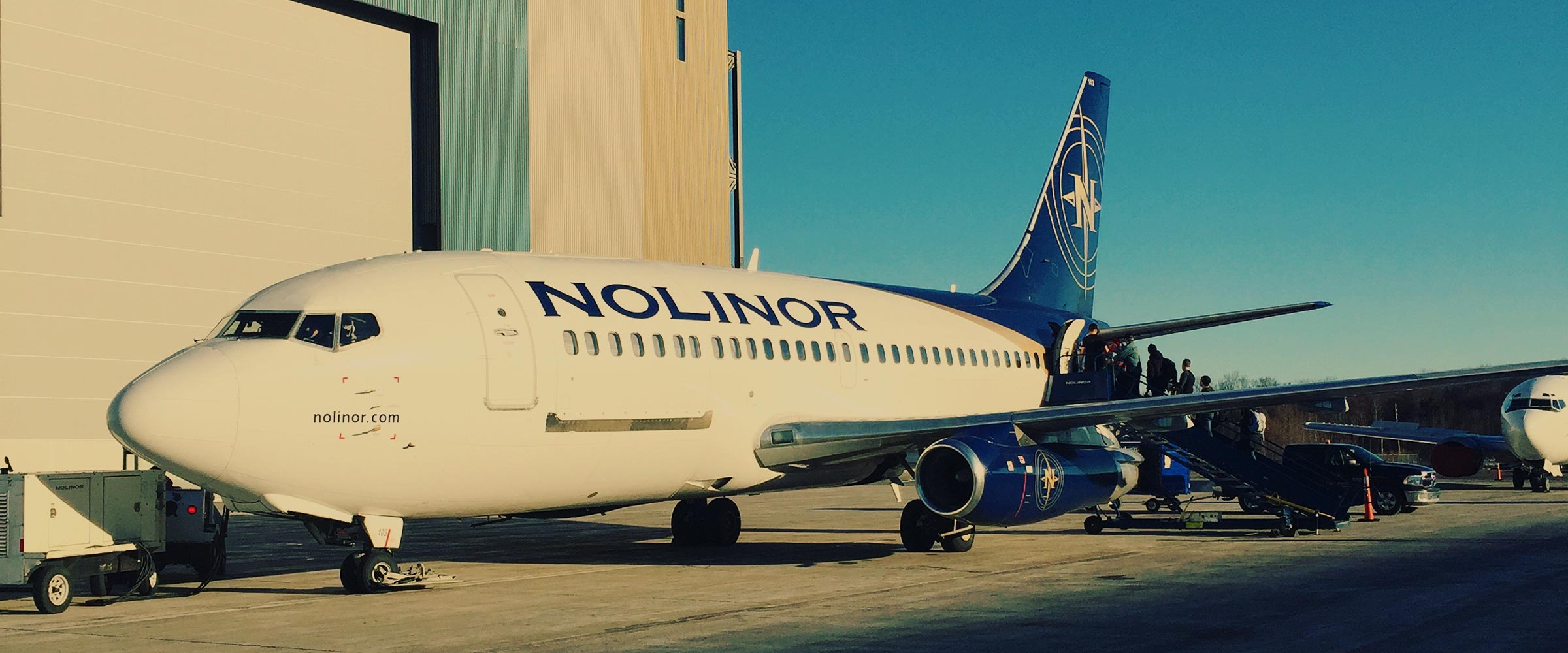 Un avion de la compagnie Nolinor est stationné sur le tarmac, des passagers montent à bord par une échelle. Le ciel est dégagé.