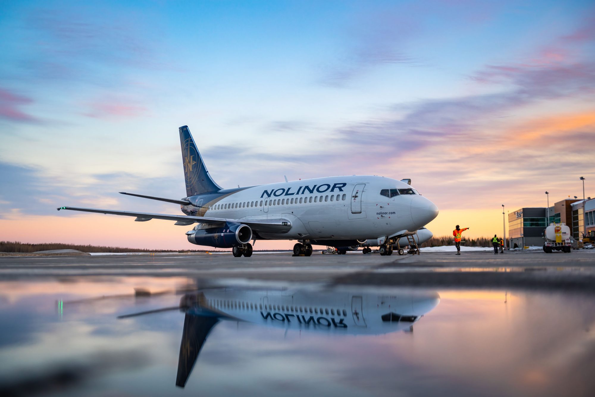 Un avion de la compagnie Nolinor est stationné sur le tarmac au crépuscule, reflété dans une flaque d'eau. Des travailleurs s'affairent à proximité.