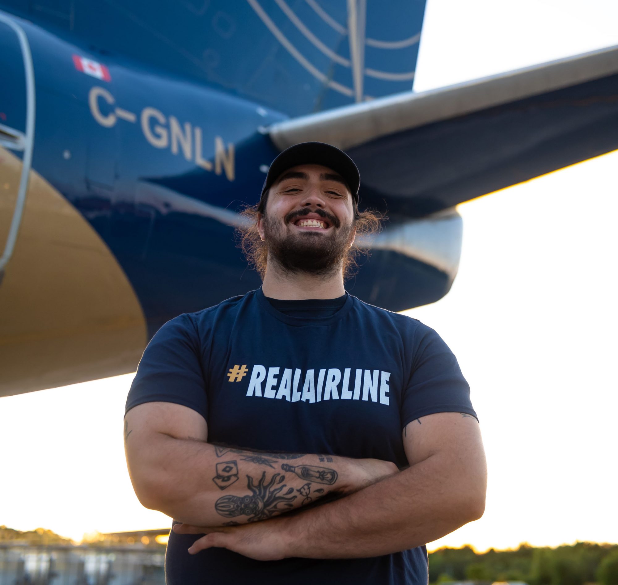 Un homme souriant porte un t-shirt avec le hashtag #REALAIRLINE, debout devant un avion aux couleurs bleu et or.