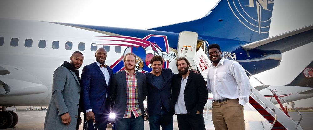 Un groupe de six hommes souriants se tient près d'un avion, portant des vêtements variés. L'avion affiche un logo coloré sur sa carlingue.