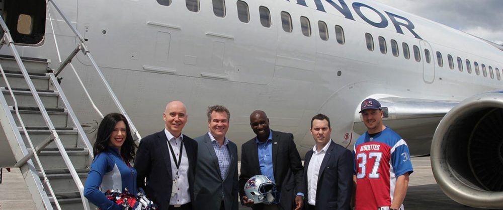 Group of six people, including cheerleaders and team members, pose with a football helmet near a plane marked "MINOR."