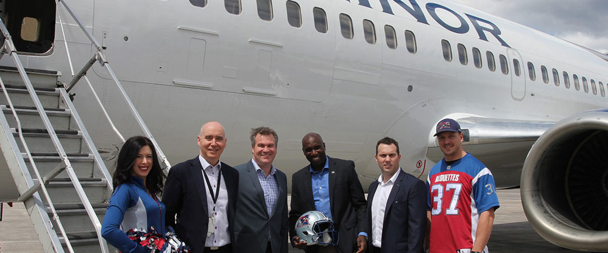 Group of six people, including cheerleaders and team members, pose with a football helmet near a plane marked "MINOR."