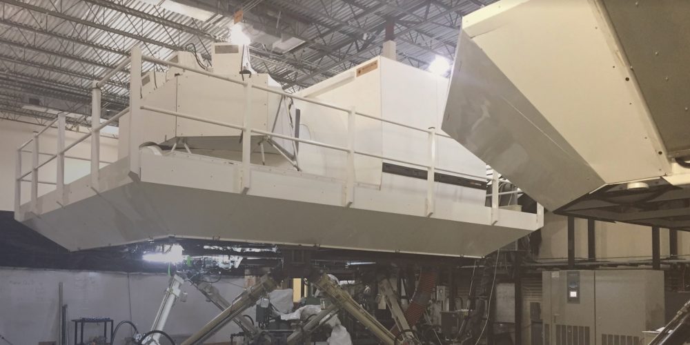 A large white structure with railings is positioned in a warehouse, connected to machinery and equipment for testing or simulation.