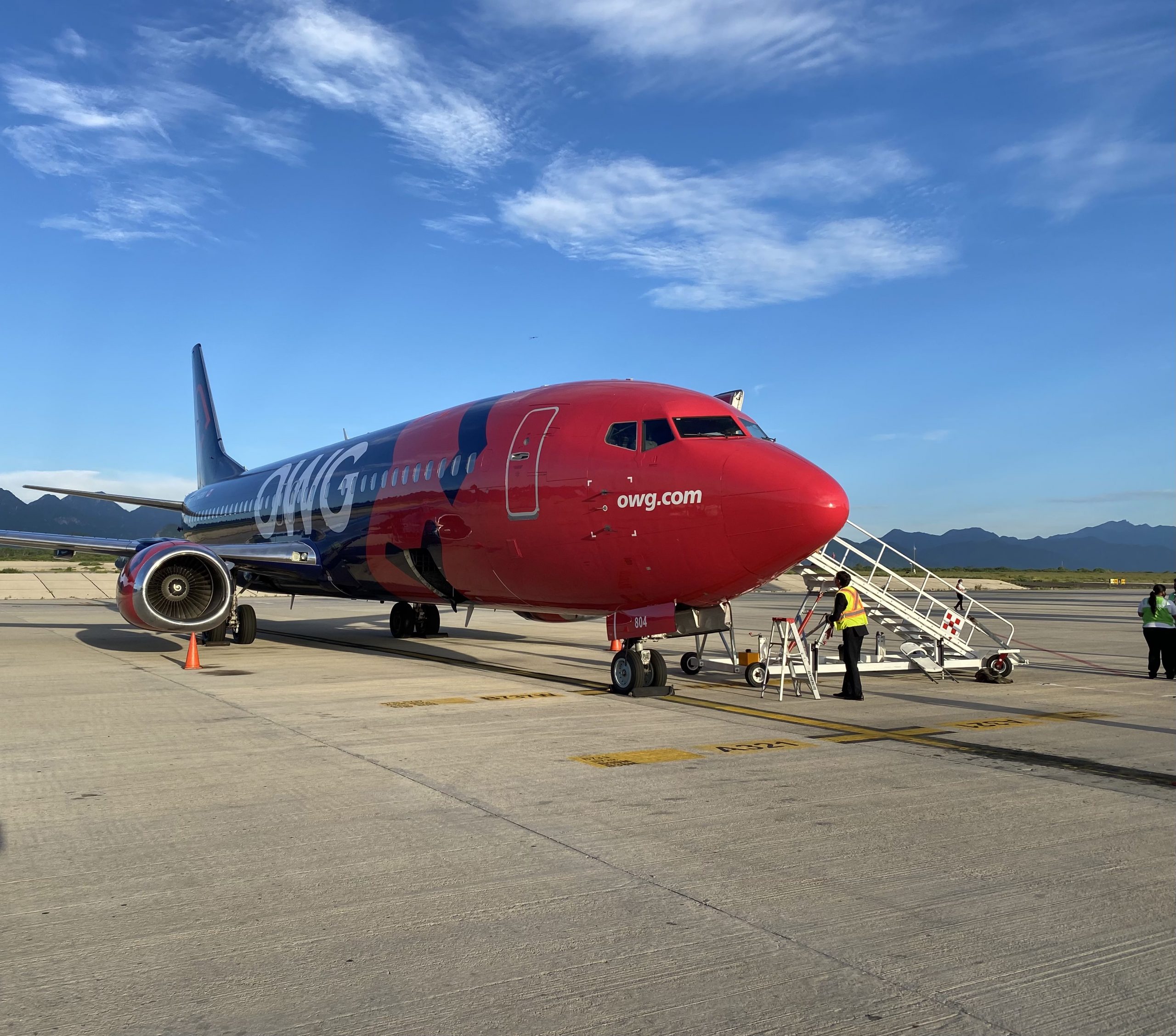 Un avion de la compagnie OWG est stationné sur le tarmac, avec des escaliers d'embarquement et des agents au sol. Le ciel est dégagé.