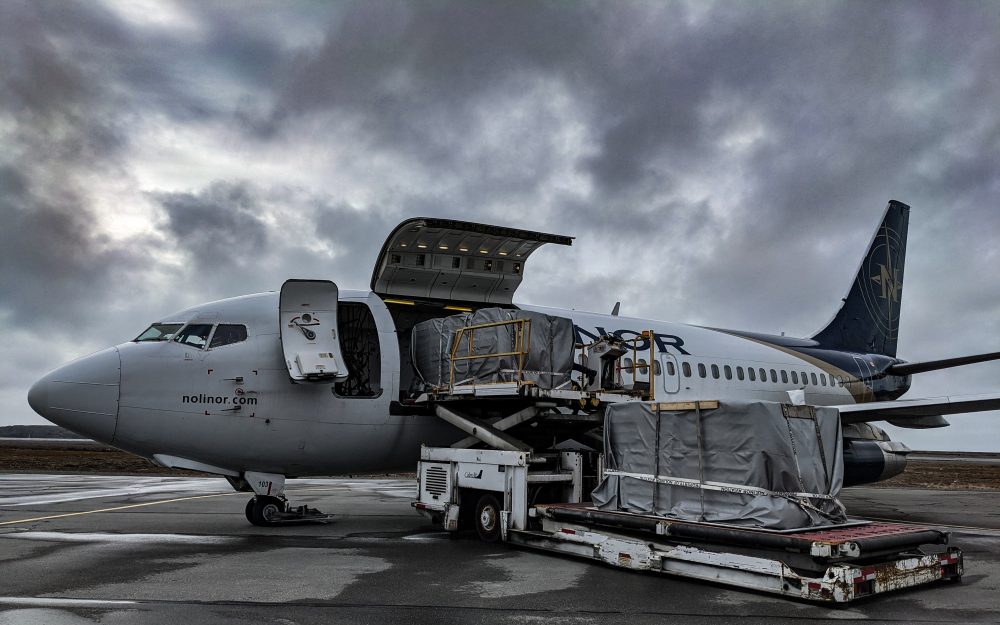 Un avion de transport de marchandises est garé sur le tarmac, avec une porte ouverte et des palettes de fret en cours de chargement. Le ciel est nuageux.