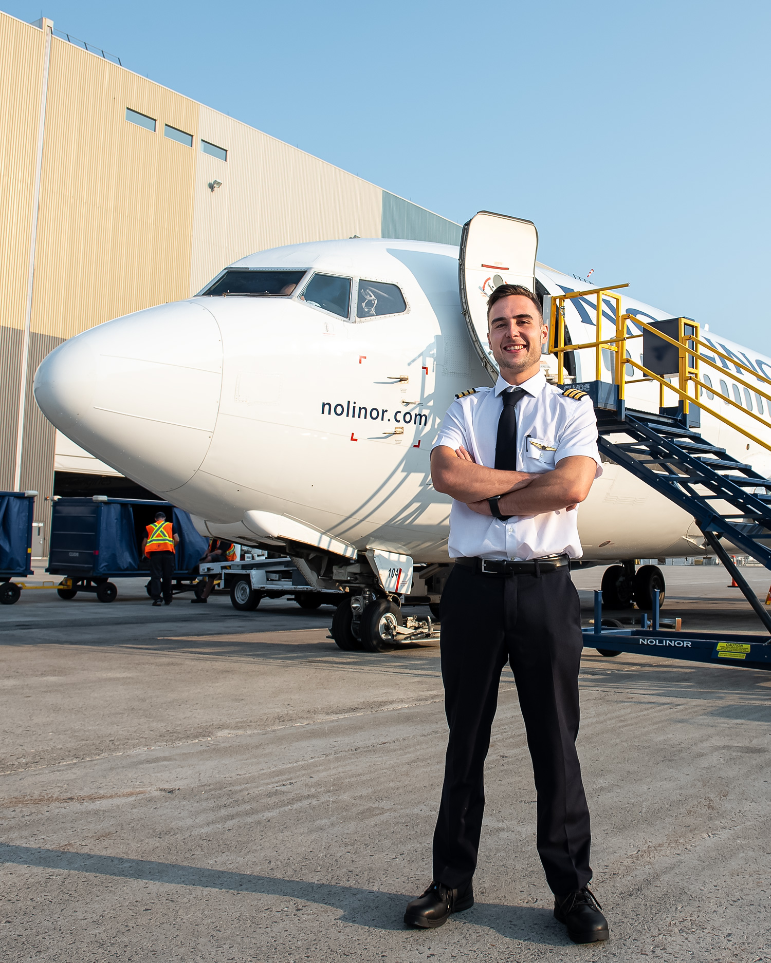 Un pilote souriant se tient devant un avion Nolinor, avec une échelle d'embarquement à proximité et un bâtiment en arrière-plan.