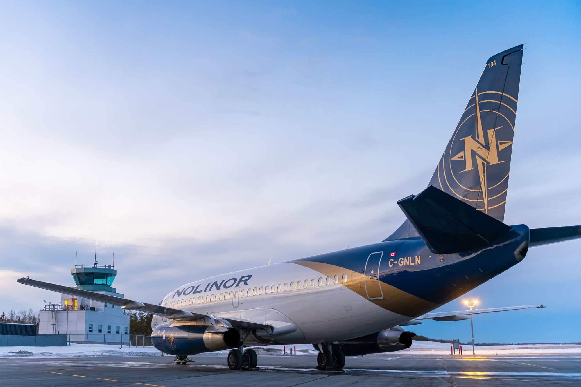 Un avion de la compagnie Nolinor est stationné sur le tarmac, avec une tour de contrôle en arrière-plan sous un ciel clair.