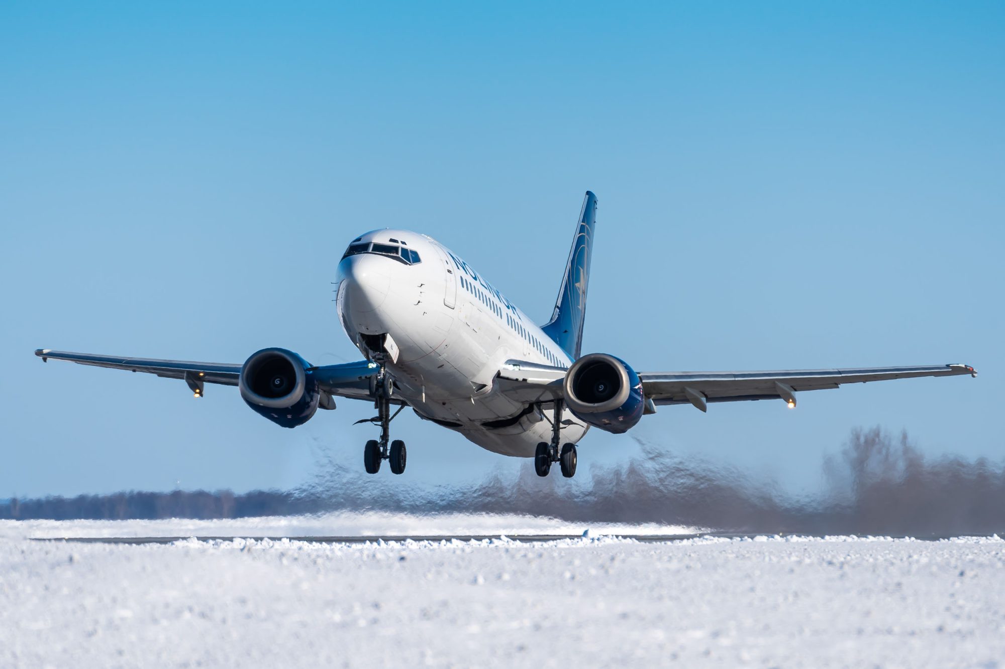 Un avion décolle d'une piste enneigée, soulevant de la neige et de la glace sous ses roues, ciel bleu en arrière-plan.