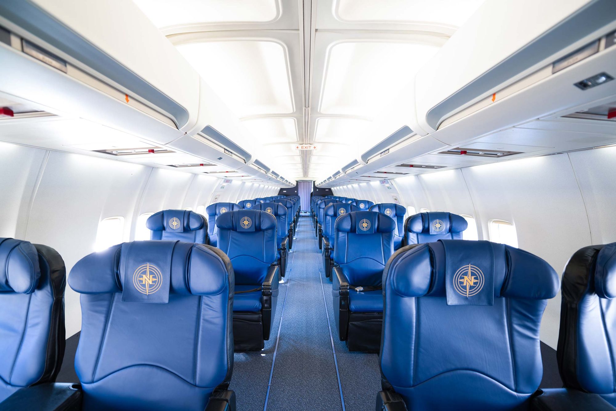 Cabine d'avion avec rangées de sièges en cuir bleu, logo sur chaque fauteuil, allée centrale dégagée et lumière naturelle.