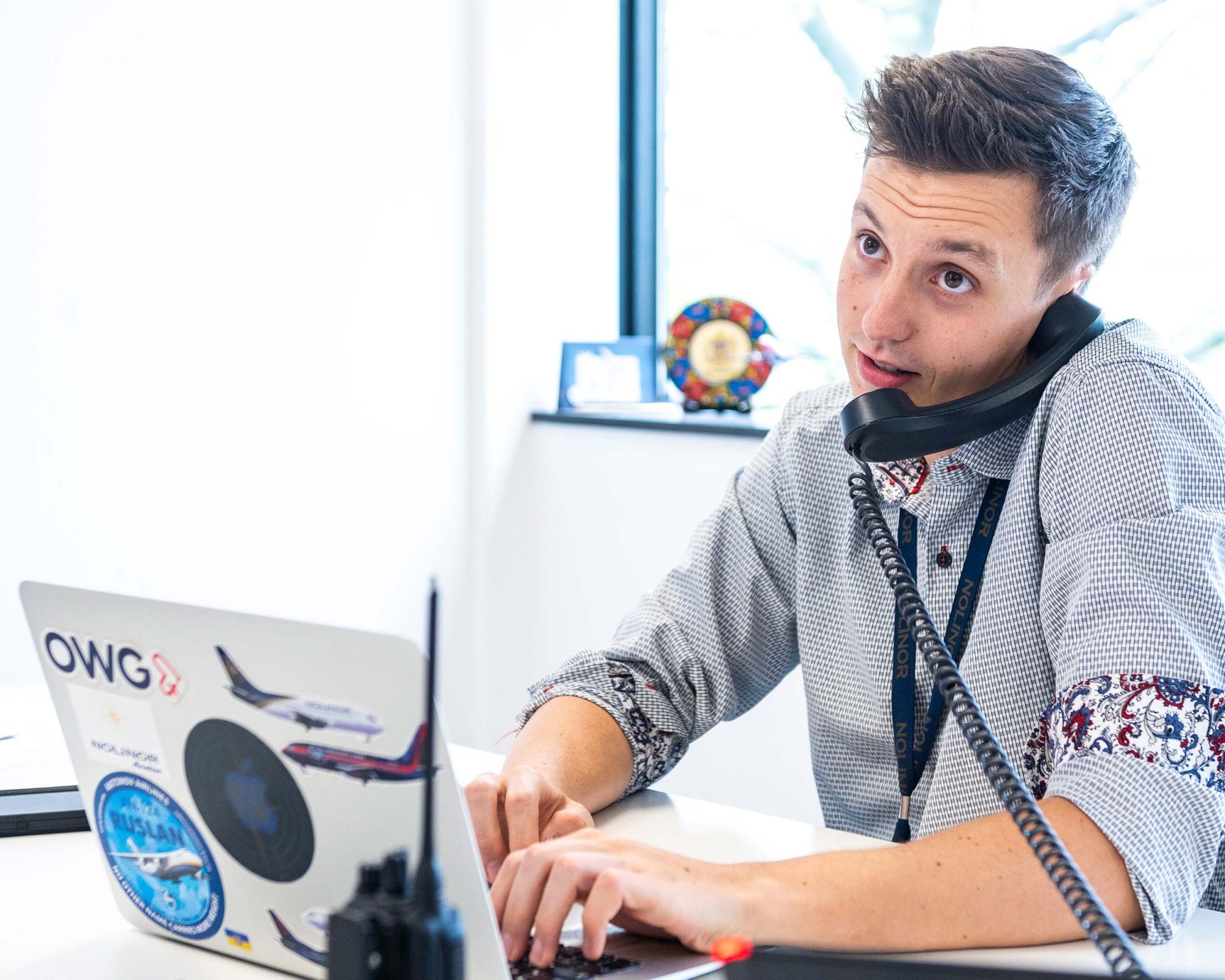Un jeune homme au bureau parle au téléphone tout en tapant sur son ordinateur portable décoré d'autocollants. Une fenêtre en arrière-plan laisse entrer la lumière.