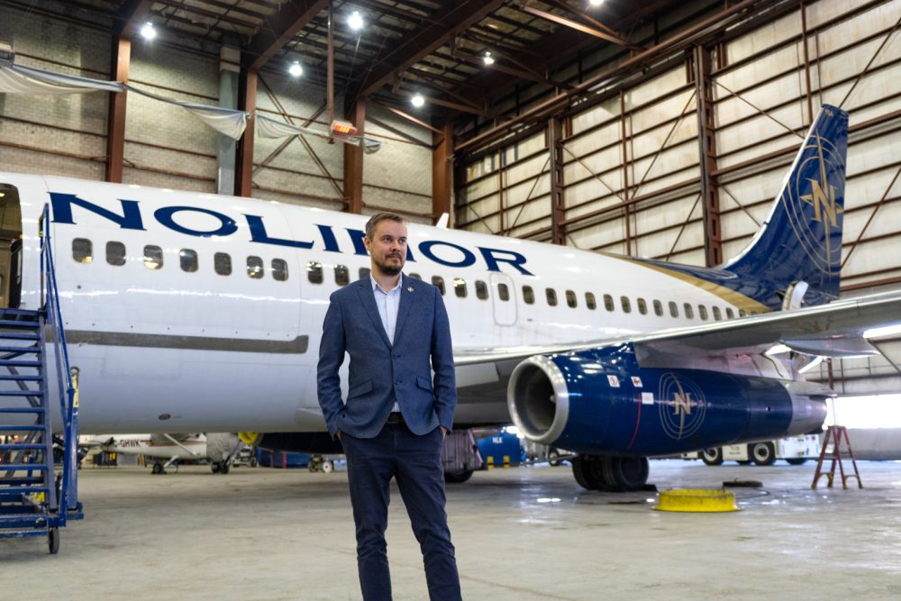 Un homme en costume se tient devant un avion de ligne dans un hangar. L'avion porte le logo de Nolitor.