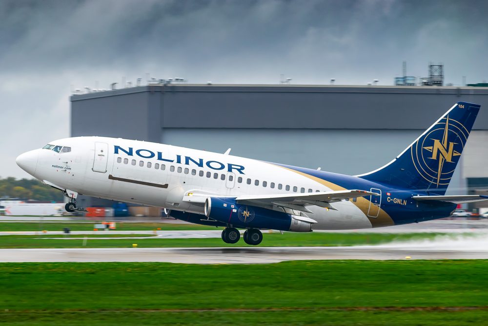 Un avion de la compagnie Nolinor décolle sur une piste humide, avec un ciel nuageux en arrière-plan. Le logo de la compagnie est bien visible sur la queue.