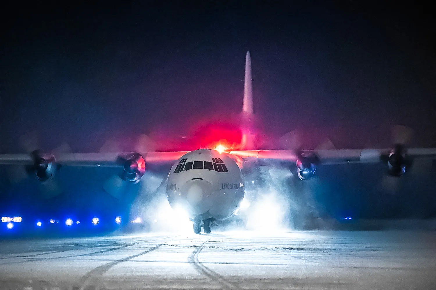 A military aircraft with lights flashing taxis on a snowy runway at night, surrounded by a misty atmosphere.