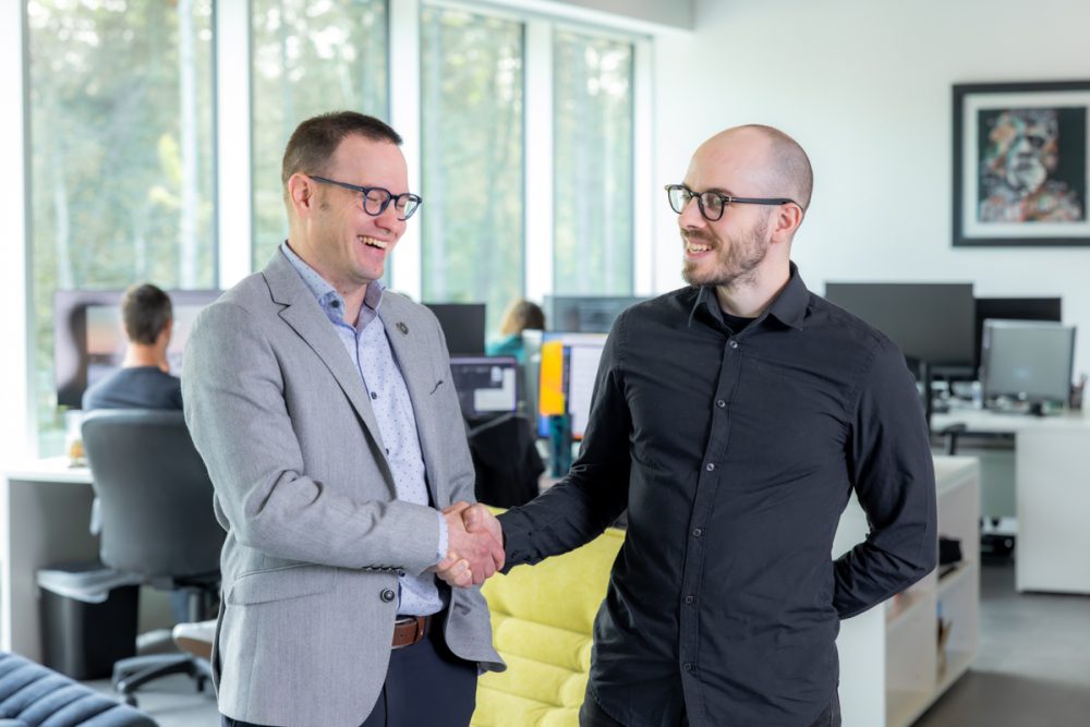 Deux hommes se serrent la main dans un bureau moderne, souriants. En arrière-plan, des collègues travaillent sur des ordinateurs.