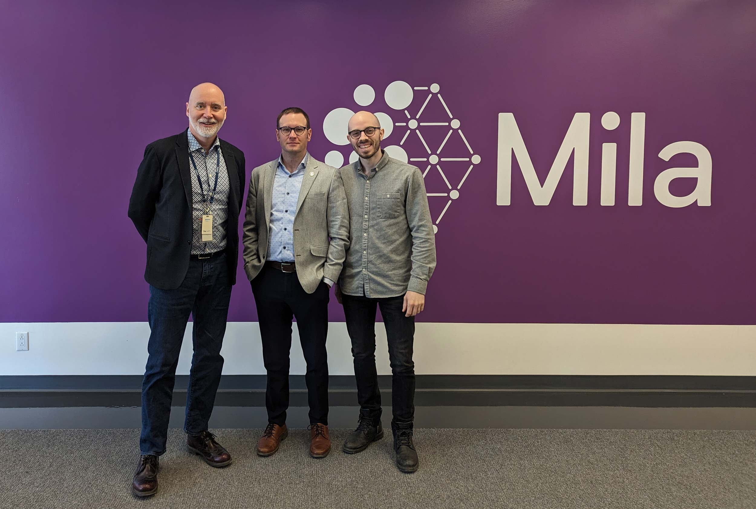 Trois hommes posent devant un mur violet avec le logo de Mila, un institut de recherche en intelligence artificielle.