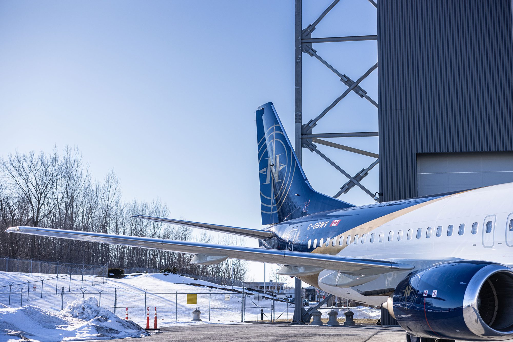 A blue and white airplane is parked near a hangar, with snow on the ground and clear skies in the background.