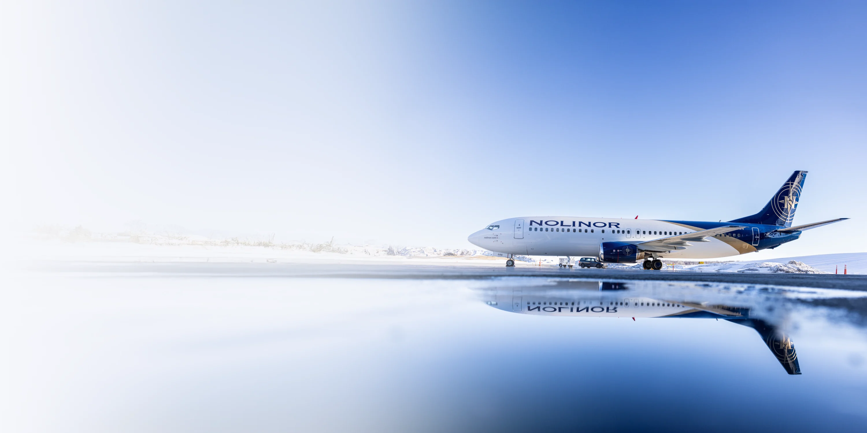 Un avion de la compagnie Nolinor est garé sur une piste, reflété dans une flaque d'eau, sous un ciel clair.