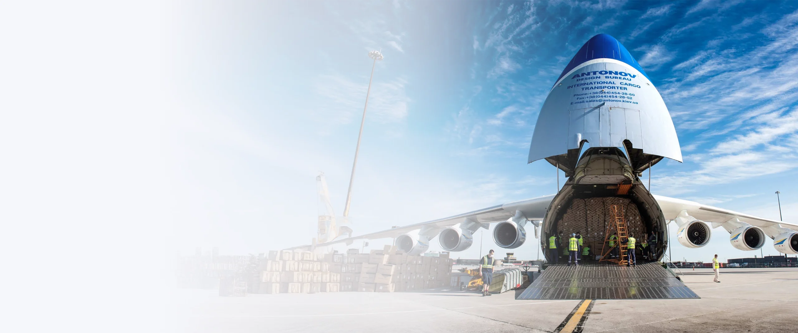 Un avion Antonov est ouvert à l'arrière, des travailleurs chargent des marchandises sur la piste sous un ciel bleu.
