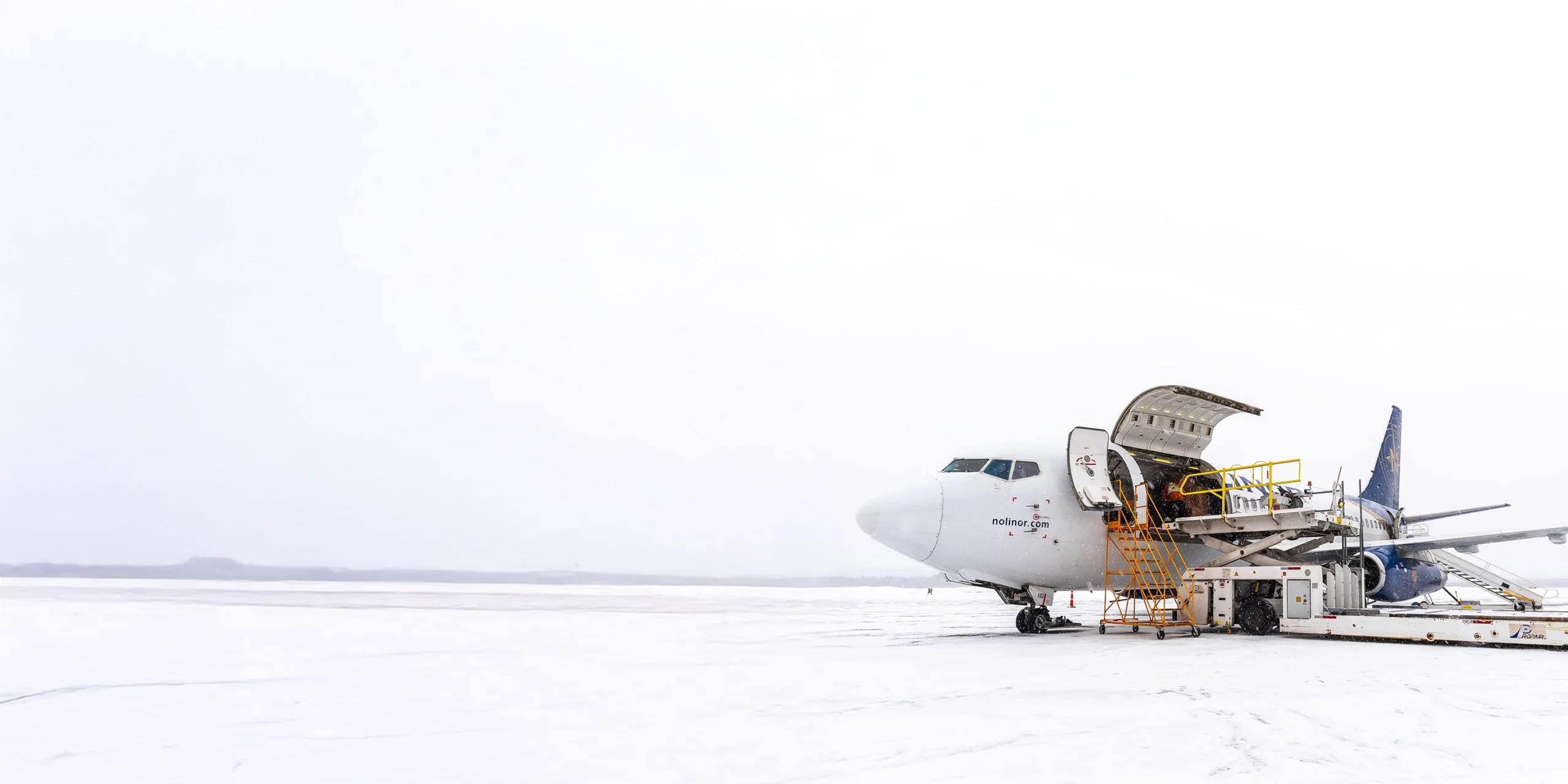A plane is parked on a snowy runway, with a service staircase attached for boarding. Snow covers the ground, indicating winter weather.