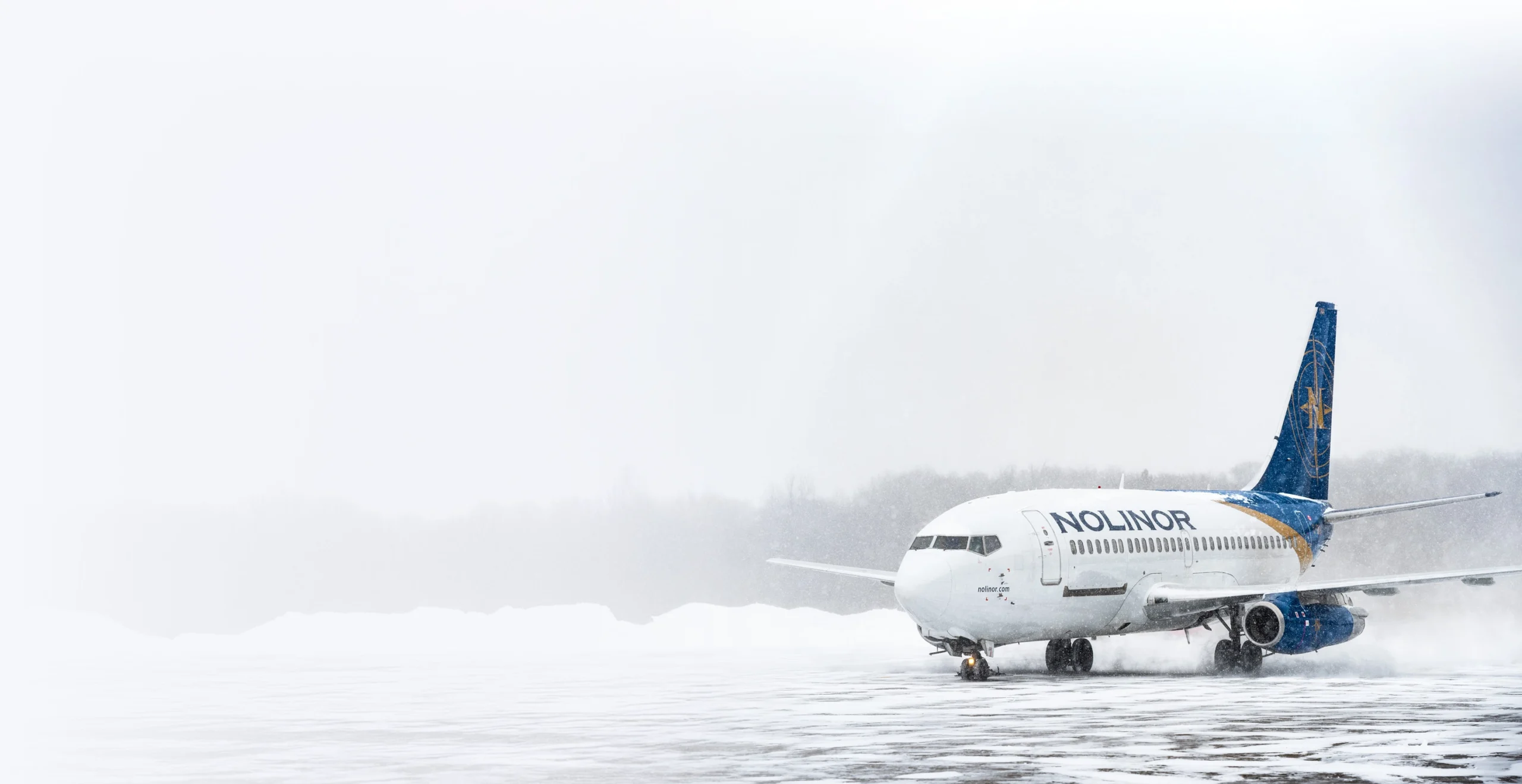 Un avion de Nolinor est stationné sur un tarmac enneigé sous une tempête de neige, créant une atmosphère hivernale.