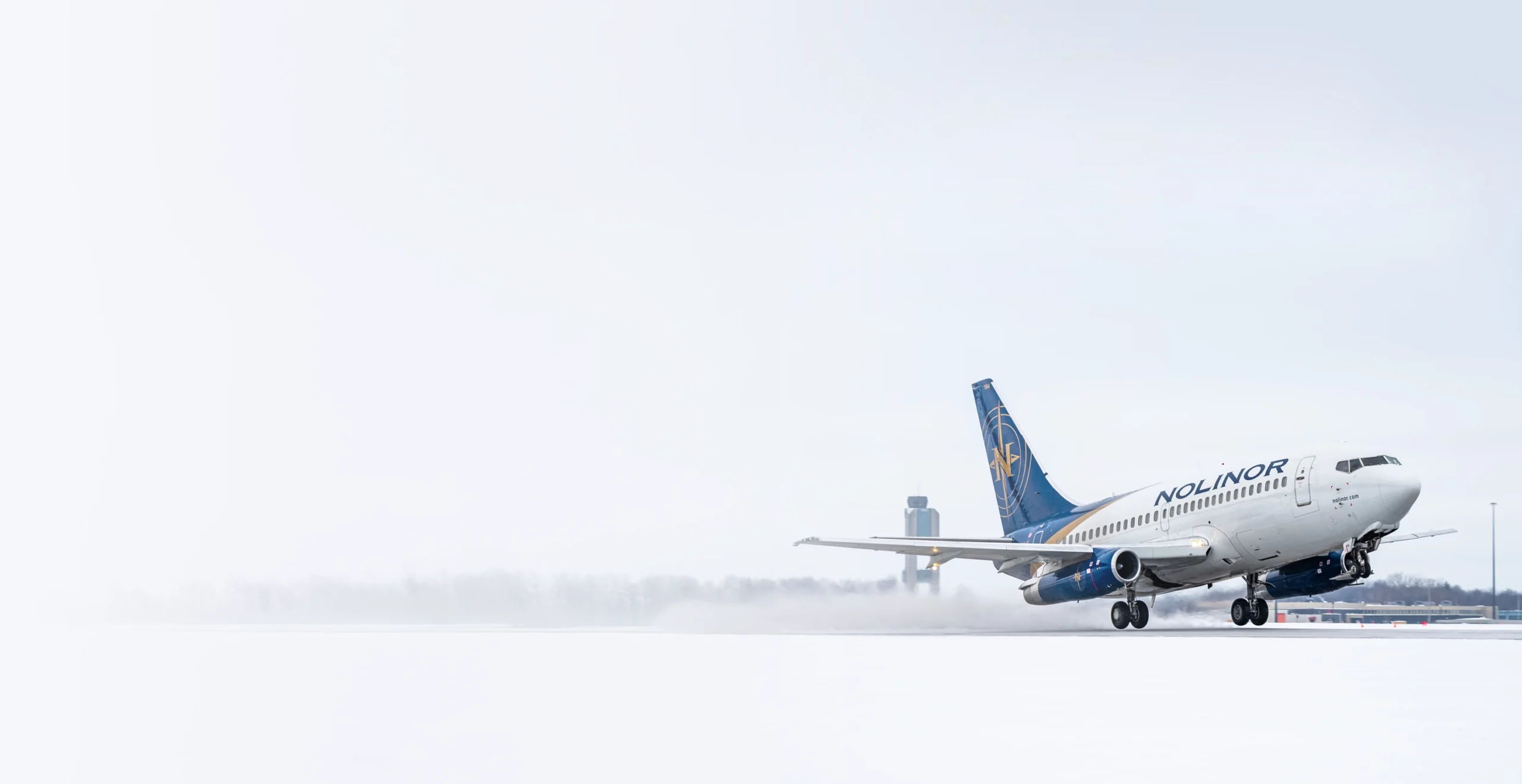 Un avion de la compagnie Nolinor décolle sur une piste enneigée, avec un aéroport en arrière-plan sous un ciel gris.