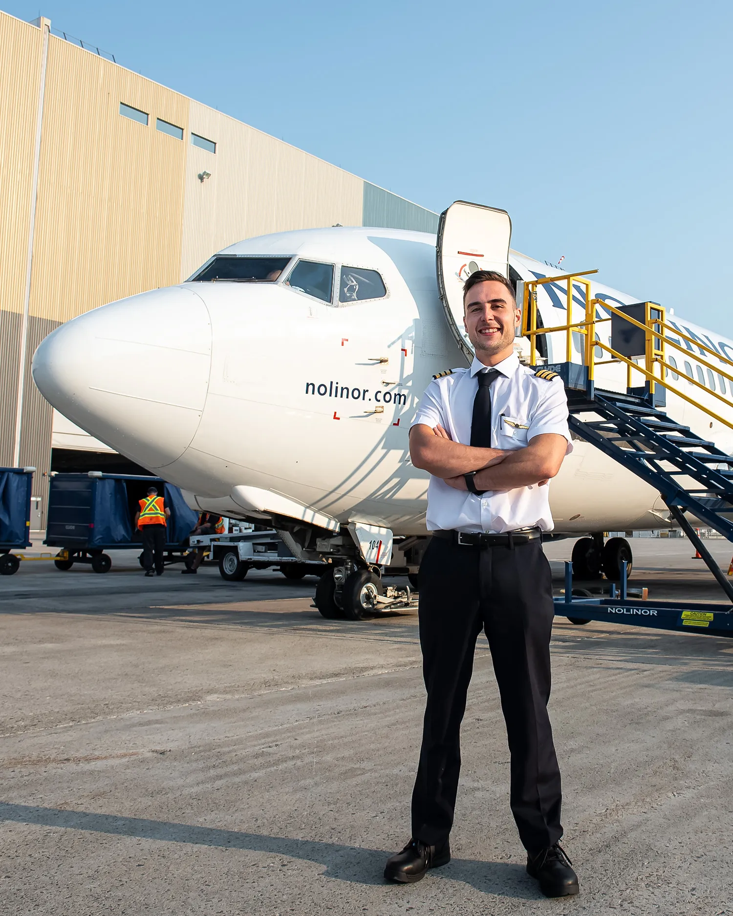 Un pilote souriant se tient devant un avion Nolinor, avec une échelle d'embarquement à proximité et un bâtiment en arrière-plan.