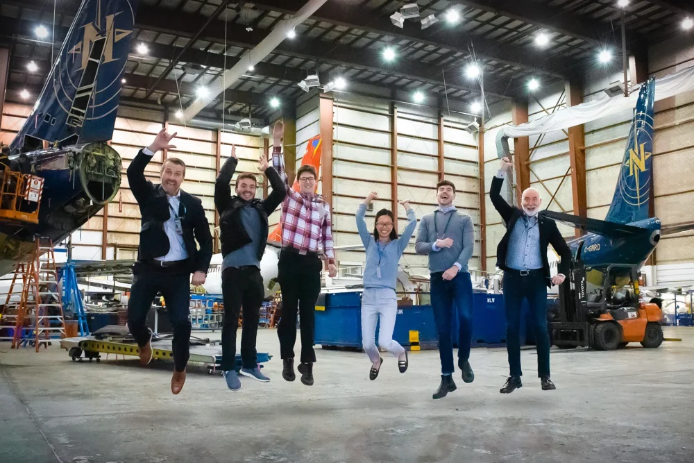 Un groupe de six personnes saute de joie dans un hangar d'avions, entouré de machines et d'équipements. L'ambiance est festive et dynamique.