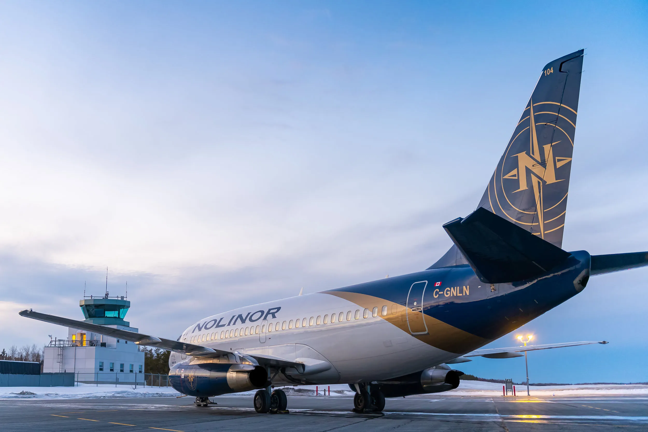Un avion de la compagnie Nolinor est stationné sur le tarmac, avec une tour de contrôle en arrière-plan sous un ciel clair.