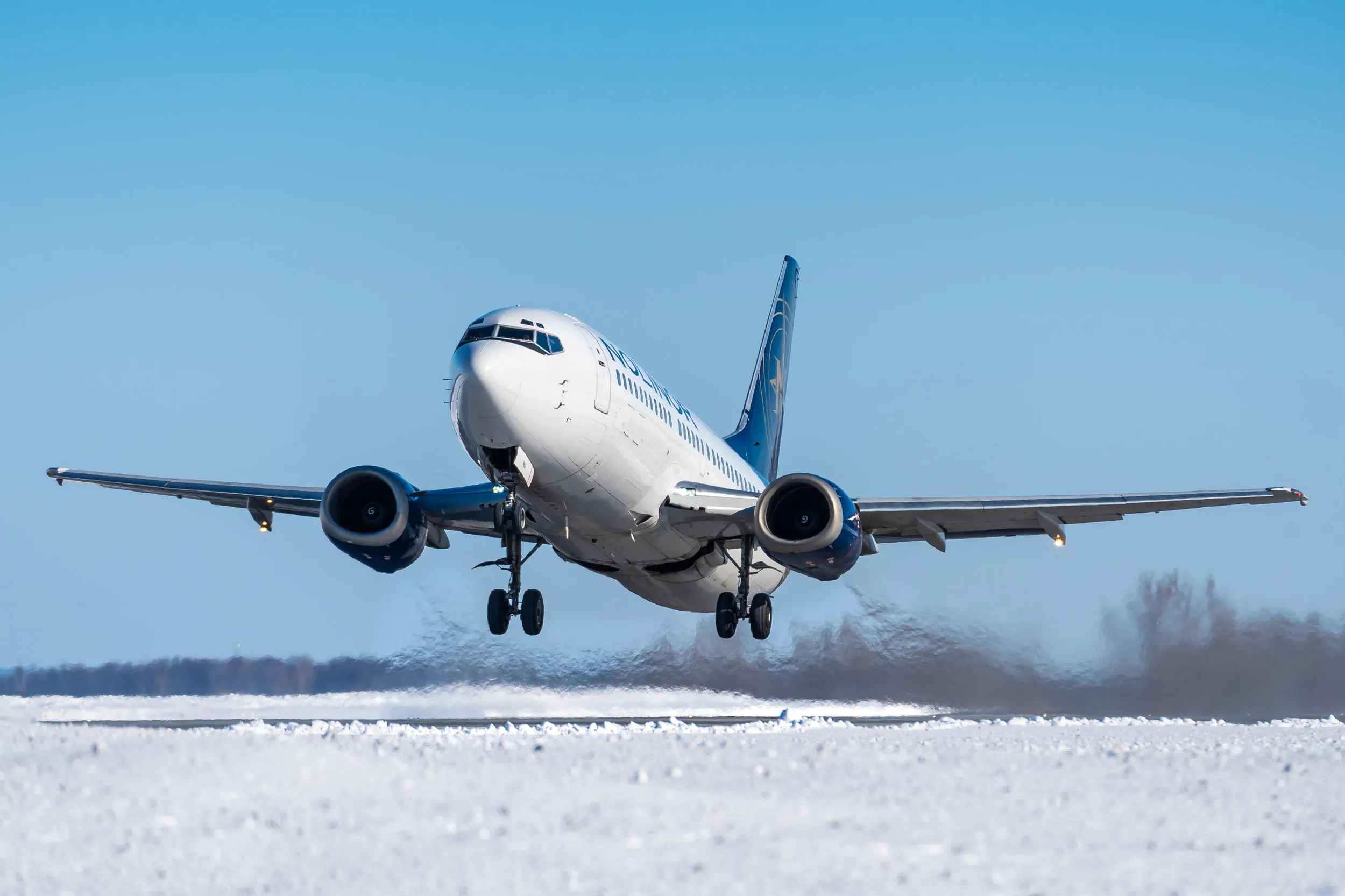 Un avion décolle d'une piste enneigée, soulevant de la neige et de la glace sous ses roues, ciel bleu en arrière-plan.
