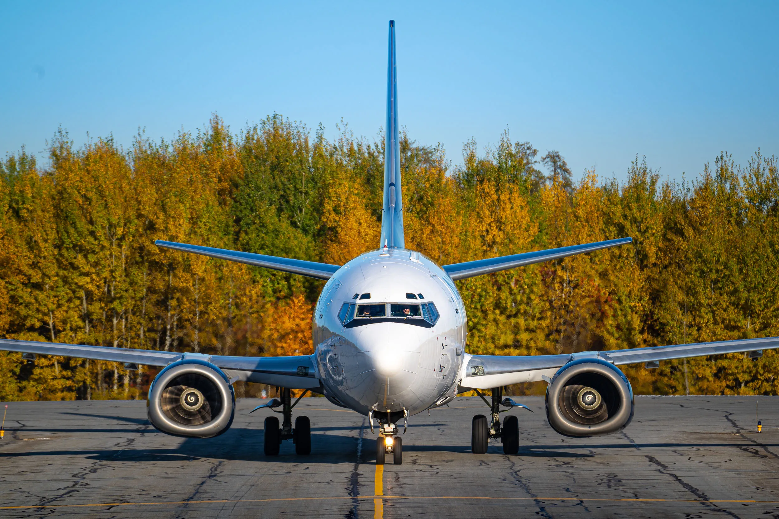 Un avion de ligne se prépare à décoller sur une piste, entouré d'arbres aux feuilles dorées en arrière-plan. Ciel clair et ensoleillé.