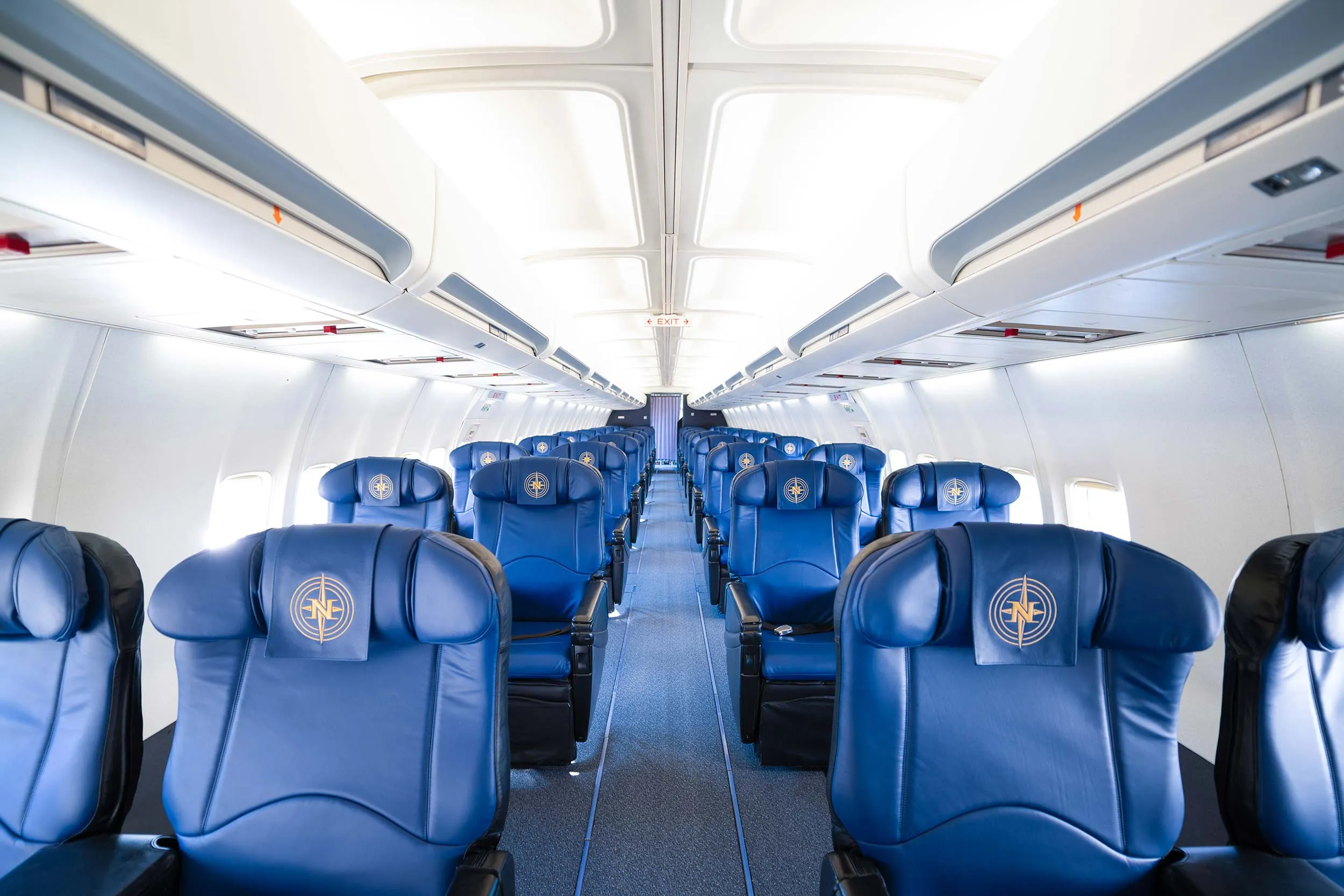 Cabine d'avion avec rangées de sièges en cuir bleu, logo sur chaque fauteuil, allée centrale dégagée et lumière naturelle.