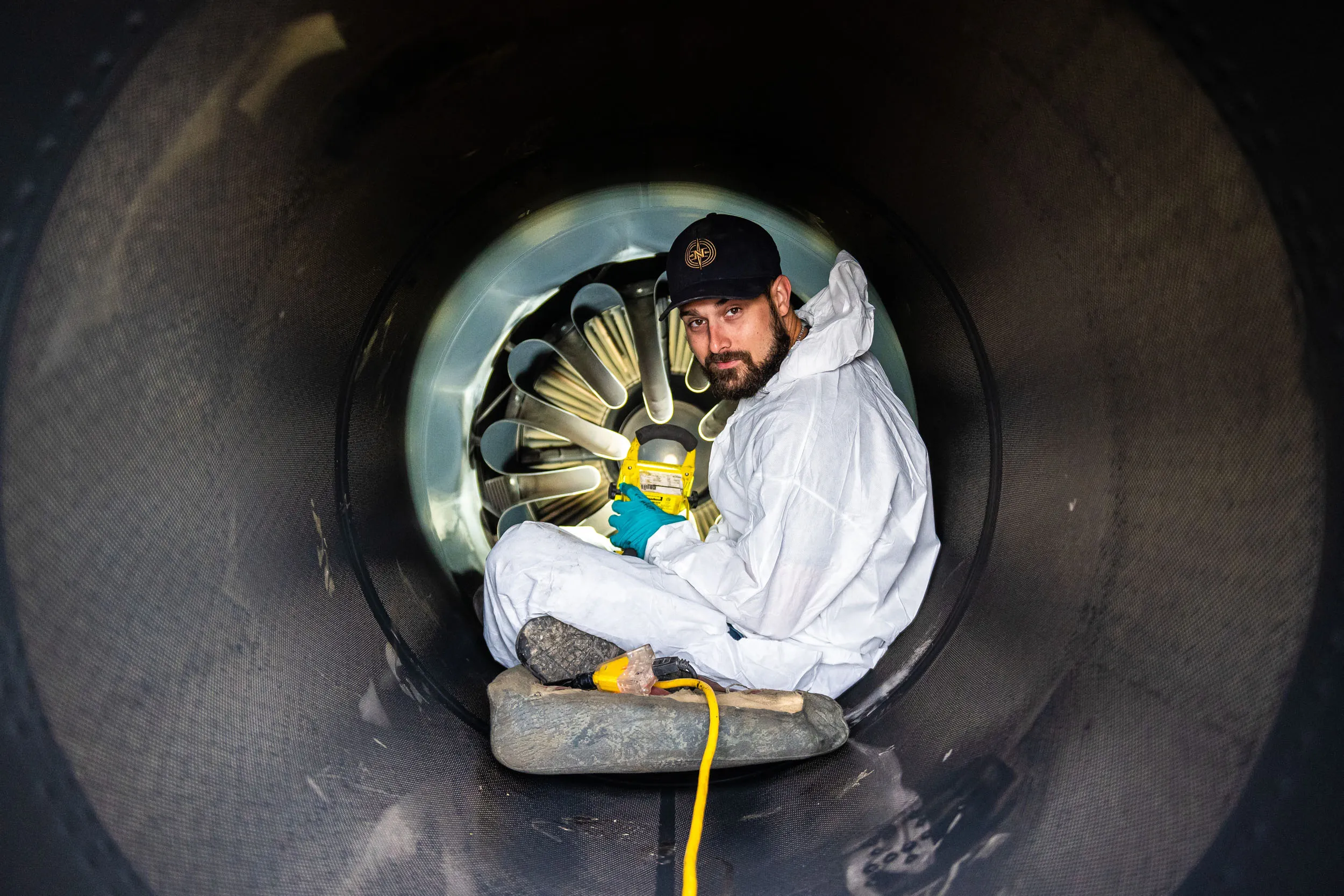 Un homme en combinaison blanche et gants bleus est assis à l'intérieur d'un grand tube noir, tenant un spray de peinture.