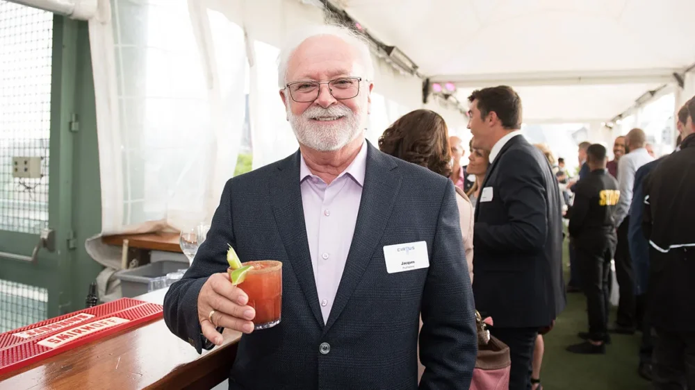 Un homme souriant tient un cocktail dans un événement en plein air, entouré de personnes en conversation. Une ambiance conviviale règne.