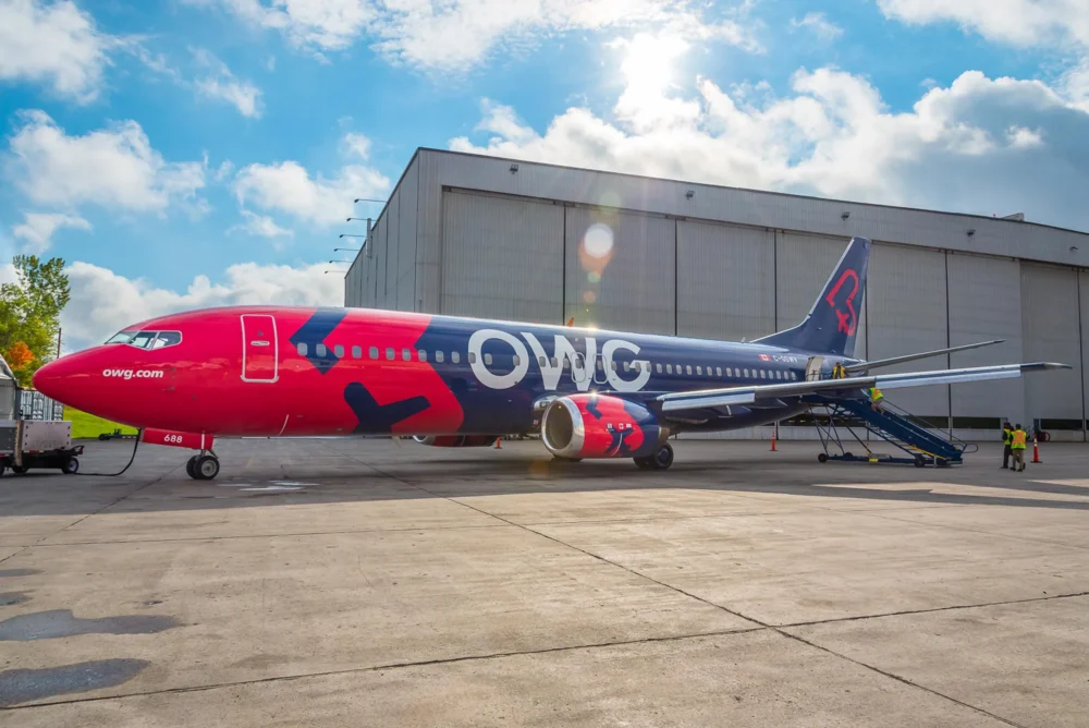A brightly colored OWG airplane parked outside a hangar, with a boarding staircase and ground crew nearby under a partly cloudy sky.
