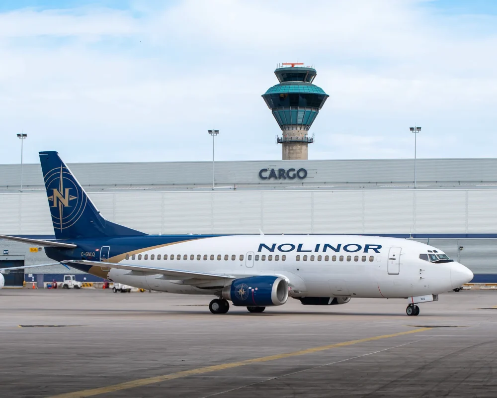 Un avion de ligne Nolinor roule sur le tarmac près d'un bâtiment de fret et d'une tour de contrôle. Le ciel est partiellement nuageux.