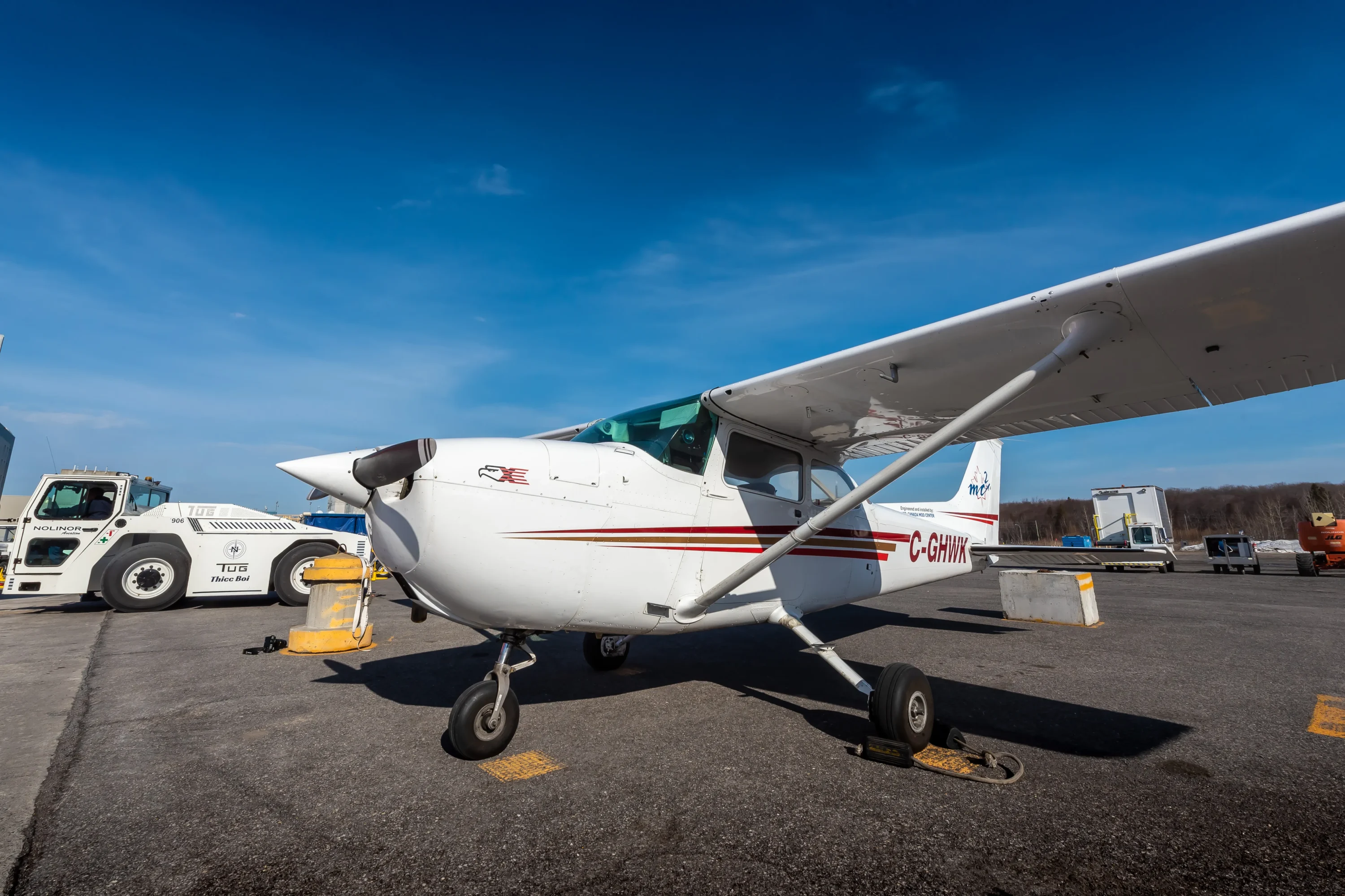 Un petit avion léger est stationné sur le tarmac sous un ciel bleu, avec un véhicule de service à proximité.