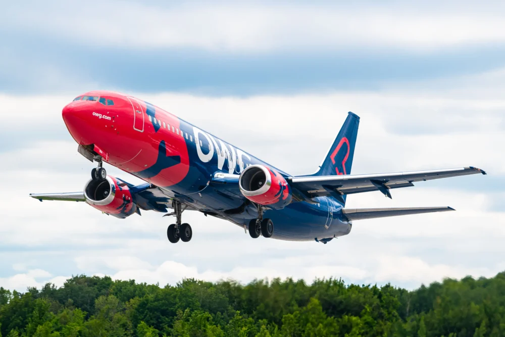 Un avion de ligne rouge et bleu décolle, avec des nuages en arrière-plan et une verdure en bas. L'appareil est en phase de montée.