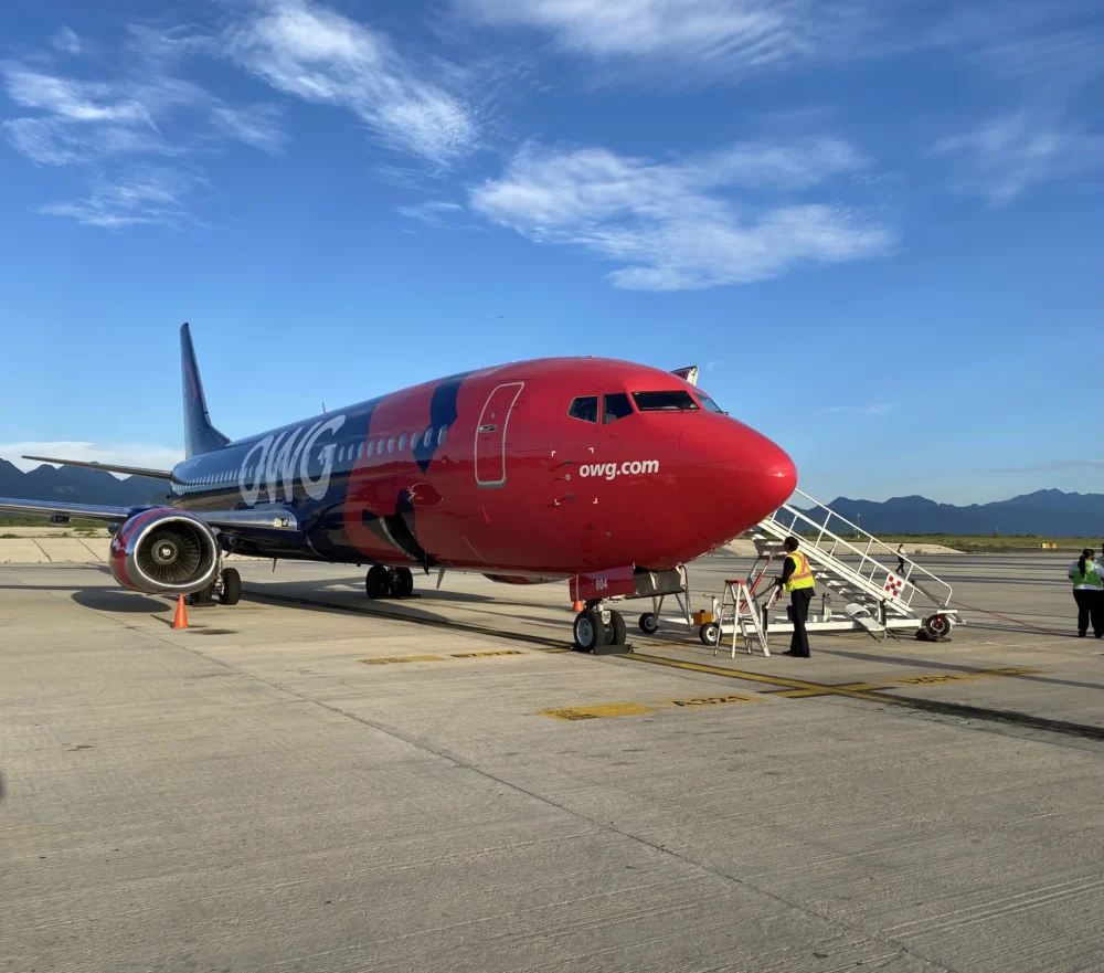 Un avion de la compagnie OWG est stationné sur le tarmac, avec des escaliers d'embarquement et des agents au sol. Le ciel est dégagé.