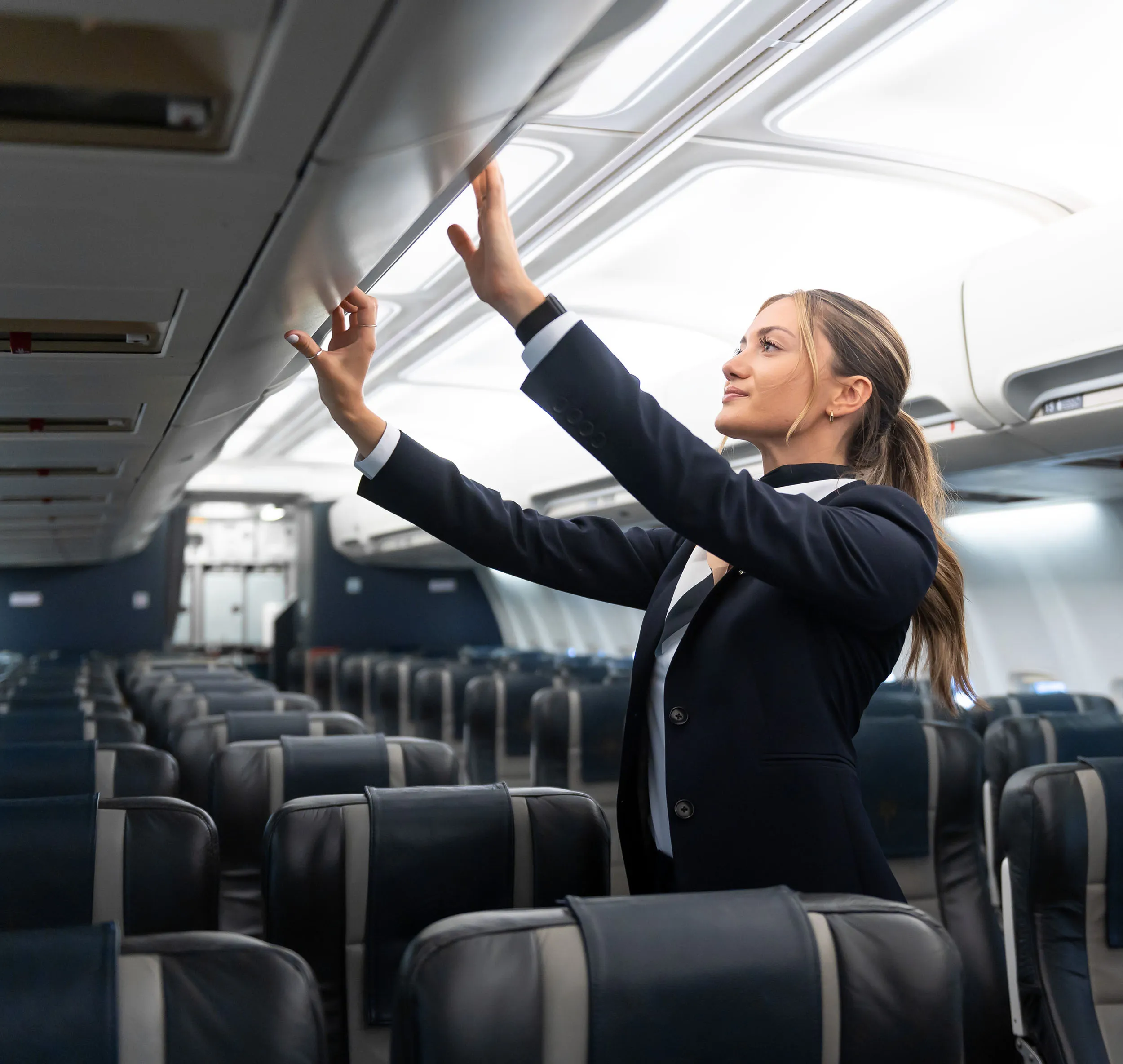 Une hôtesse de l'air ajuste le compartiment à bagages dans un avion, avec des sièges vides en arrière-plan. Elle porte un uniforme professionnel.