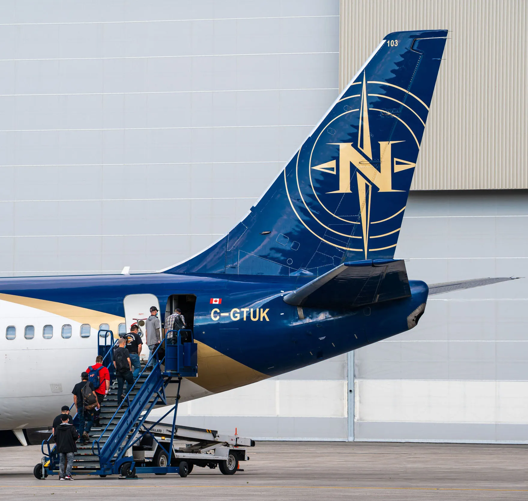 Des passagers montent à bord d'un avion, le C-GTUK, par une échelle mobile. L'arrière de l'appareil présente un design bleu et or.
