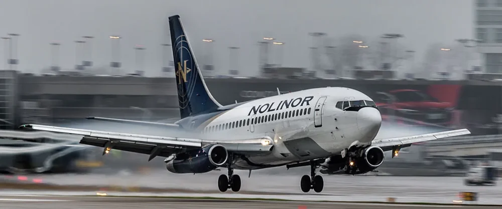 Un avion de la compagnie Nolinor atterrit sur une piste mouillée, avec des lumières d'aéroport en arrière-plan. Le ciel est nuageux.