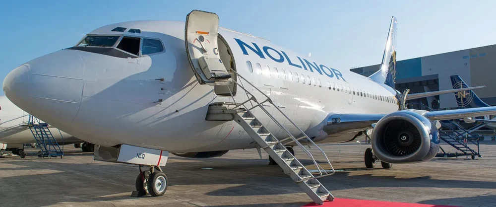 Un avion de la compagnie Nolinor est stationné sur le tarmac, avec une échelle d'embarquement à l'avant et un ciel dégagé en arrière-plan.