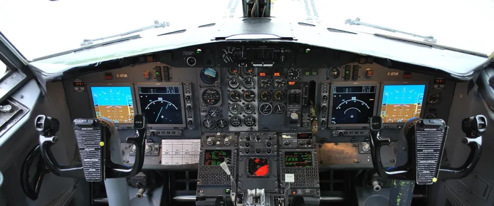Poste de pilotage d'un avion avec plusieurs écrans affichant des instruments de vol et des commandes. Les manettes et boutons sont bien visibles.