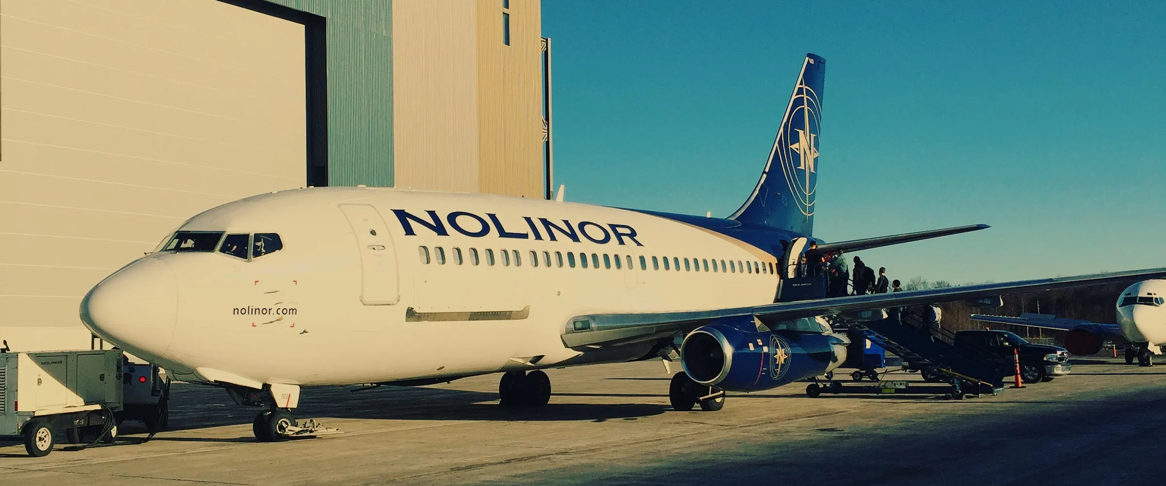Un avion de la compagnie Nolinor est stationné sur le tarmac, des passagers montent à bord par une échelle. Le ciel est dégagé.
